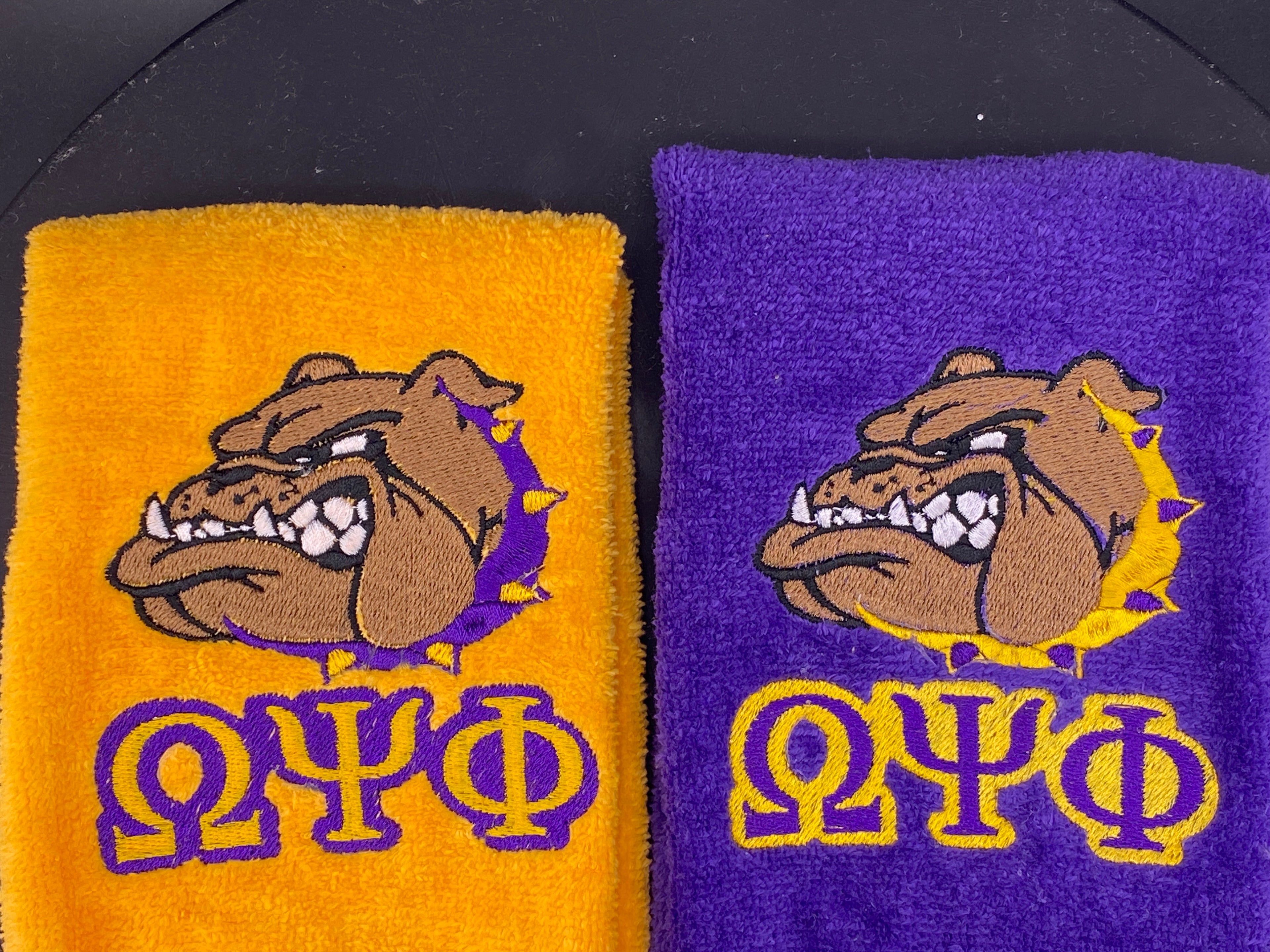 2 plush velour Leisure Luxe Towels embroidered with the iconic Bulldawg and greek letters. One towel is gold with a purple collar on the dawg, the other is purple with a gold collar on the dawg. 